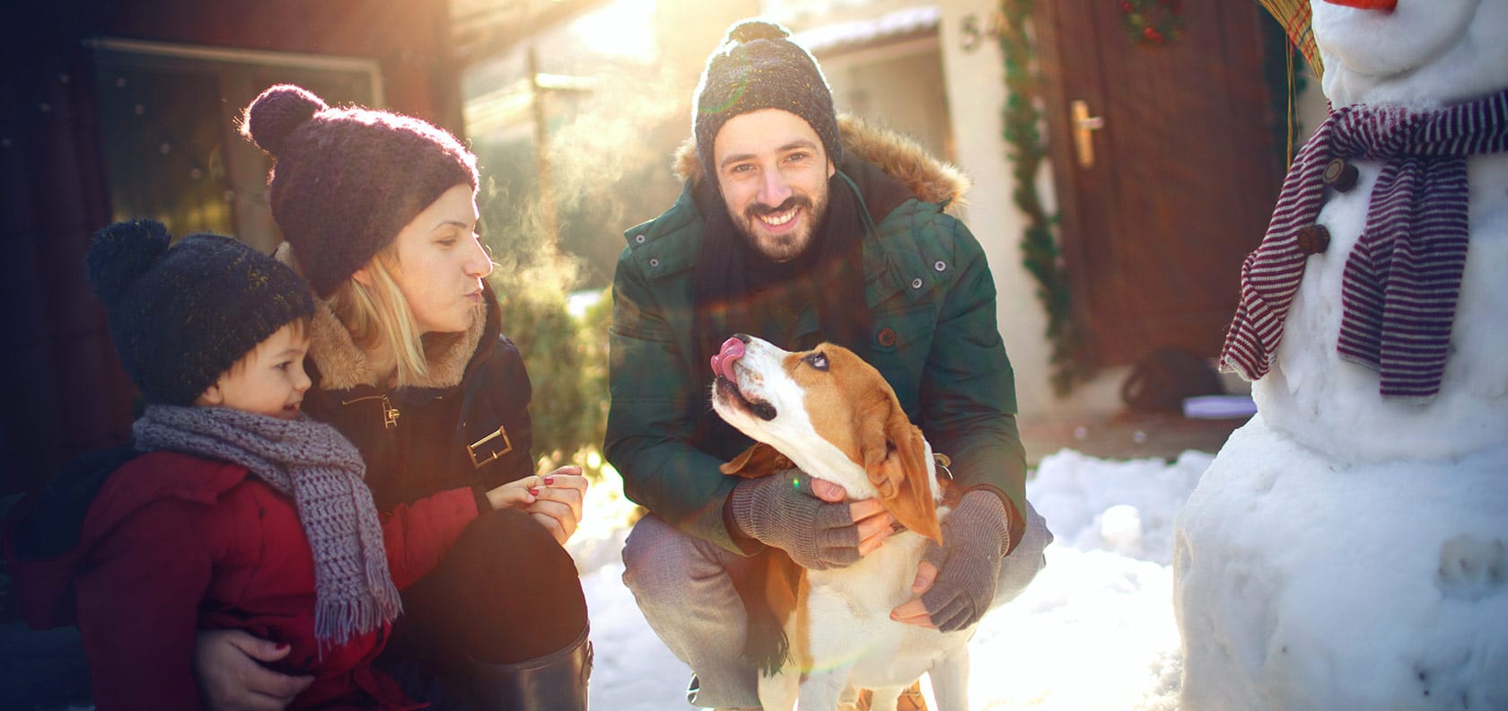 Family dressed in winter clothes with their pet in the courtyard of their house building a snowman.